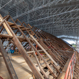 Das Bild zeigt eine große Baustelle im Inneren eines Gebäudes mit umfangreichem Metallgerüst und einem hölzernen Dachstuhl aus mehreren dreieckigen Trägern und Balken. Leitern und Baumaterialien deuten auf laufende Arbeiten hin. Eine schützende Metallüberdachung überspannt den gesamten Bereich, was auf die Sanierung eines historischen Gebäudes schließen lässt.
