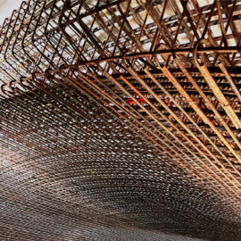 Das Bild zeigt eine Nahaufnahme einer komplexen Bewehrungskonstruktion aus Stahlstäben, die für den Tunnelbau verwendet wird. Die Stäbe sind eng miteinander verflochten und bilden ein dichtes Gitter, das sich bogenförmig nach unten krümmt. Im Hintergrund sind weitere Bauelemente und Rohrleitungen zu erkennen, die die technische Umgebung der Baustelle verdeutlichen. Die gesamte Szene ist gut ausgeleuchtet und hebt die Struktur und Textur des rostfarbenen Stahls hervor.