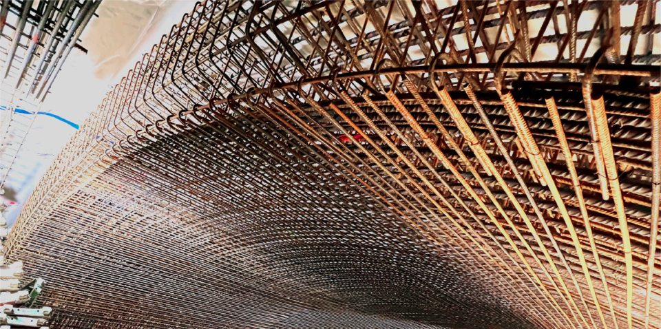 Das Bild zeigt eine Nahaufnahme einer komplexen Bewehrungskonstruktion aus Stahlstäben, die für den Tunnelbau verwendet wird. Die Stäbe sind eng miteinander verflochten und bilden ein dichtes Gitter, das sich bogenförmig nach unten krümmt. Im Hintergrund sind weitere Bauelemente und Rohrleitungen zu erkennen, die die technische Umgebung der Baustelle verdeutlichen. Die gesamte Szene ist gut ausgeleuchtet und hebt die Struktur und Textur des rostfarbenen Stahls hervor.