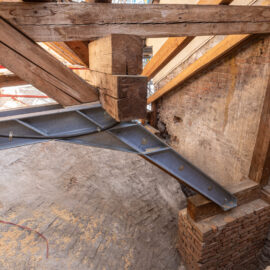 Das Bild zeigt eine große Baustelle im Inneren eines Gebäudes mit umfangreichem Metallgerüst und einem hölzernen Dachstuhl aus mehreren dreieckigen Trägern und Balken. Leitern und Baumaterialien deuten auf laufende Arbeiten hin. Eine schützende Metallüberdachung überspannt den gesamten Bereich, was auf die Sanierung eines historischen Gebäudes schließen lässt.