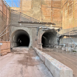 Das Bild zeigt den Eingang zu zwei parallel verlaufenden Tunnelröhren, die für den U-Bahn-Bau vorgesehen sind. Die Tunnelwände bestehen aus grobem Spritzbeton, und an den Öffnungen sind Kabel und Leitungen befestigt. Der Boden vor den Tunnelportalen ist schlammig und teilweise mit Wasser bedeckt, während eine massive Betonbarriere den Zugang teilt. Im oberen Bereich sind Holzverschalungen und Stützkonstruktionen sichtbar, die den Bauabschnitt absichern.