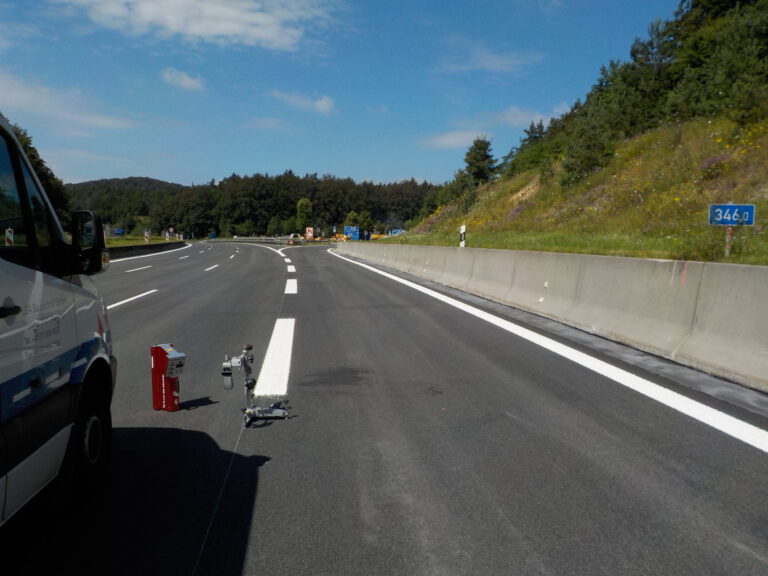 Das Bild zeigt einen Abschnitt einer Autobahn mit frisch aufgetragenen weißen Fahrbahnmarkierungen. Auf der linken Seite ist ein Fahrzeug teilweise zu sehen, daneben steht Ausrüstung, vermutlich für Markierungsarbeiten. Die Straße verläuft in einer Rechtskurve und ist von Bäumen und Grünflächen umgeben.