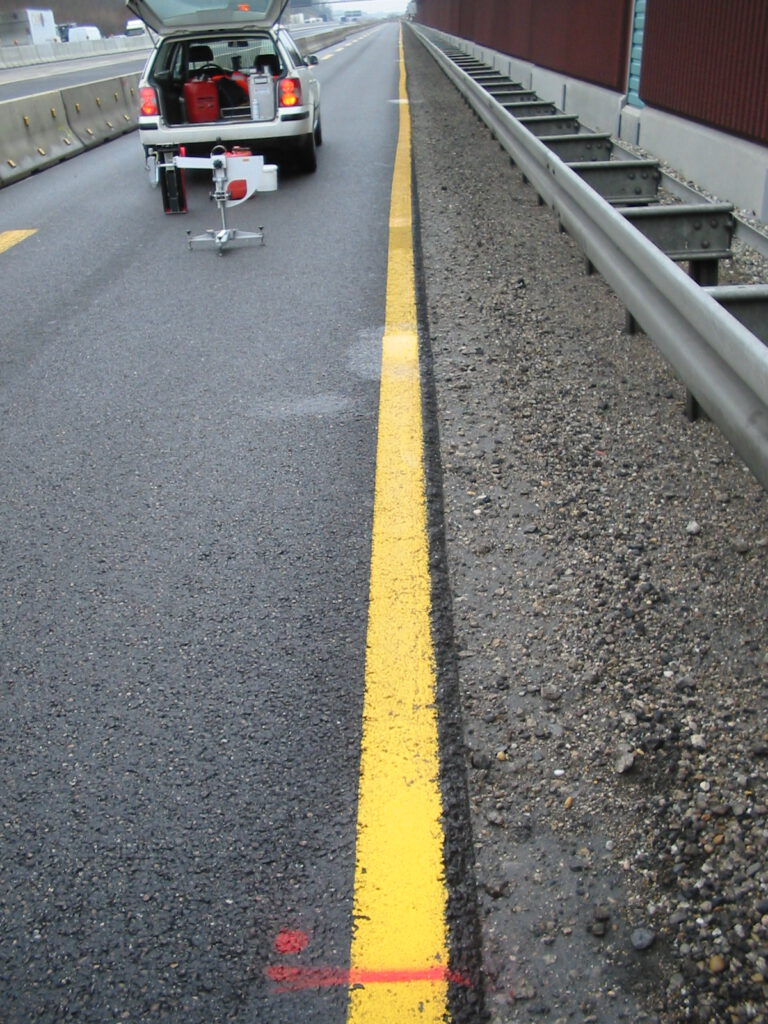 Das Bild zeigt einen Abschnitt einer Straße mit einer gelben Linie am Fahrbahnrand. Auf der linken Seite steht ein Auto mit geöffnetem Kofferraum, an dem hinten Gerät befestigt ist, für Wartungs- oder Vermessungsarbeiten. Rechts neben der Straße befinden sich ein Leitplanke und ein Schotterstreifen.