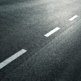 Das Bild zeigt einen Abschnitt einer Asphaltstraße mit einer gestrichelten weißen Linie in der Mitte. Die Lichtverhältnisse deuten auf frühen Morgen oder späten Nachmittag hin, erkennbar an den Schatten und Lichtreflexionen auf dem Belag. Es sind keine Fahrzeuge oder weiteren Objekte zu sehen.