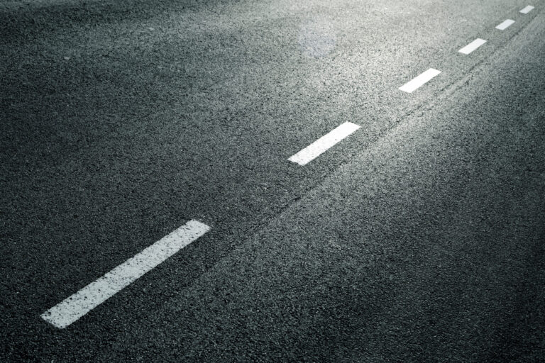 Das Bild zeigt einen Abschnitt einer Asphaltstraße mit einer gestrichelten weißen Linie in der Mitte. Die Lichtverhältnisse deuten auf frühen Morgen oder späten Nachmittag hin, erkennbar an den Schatten und Lichtreflexionen auf dem Belag. Es sind keine Fahrzeuge oder weiteren Objekte zu sehen.