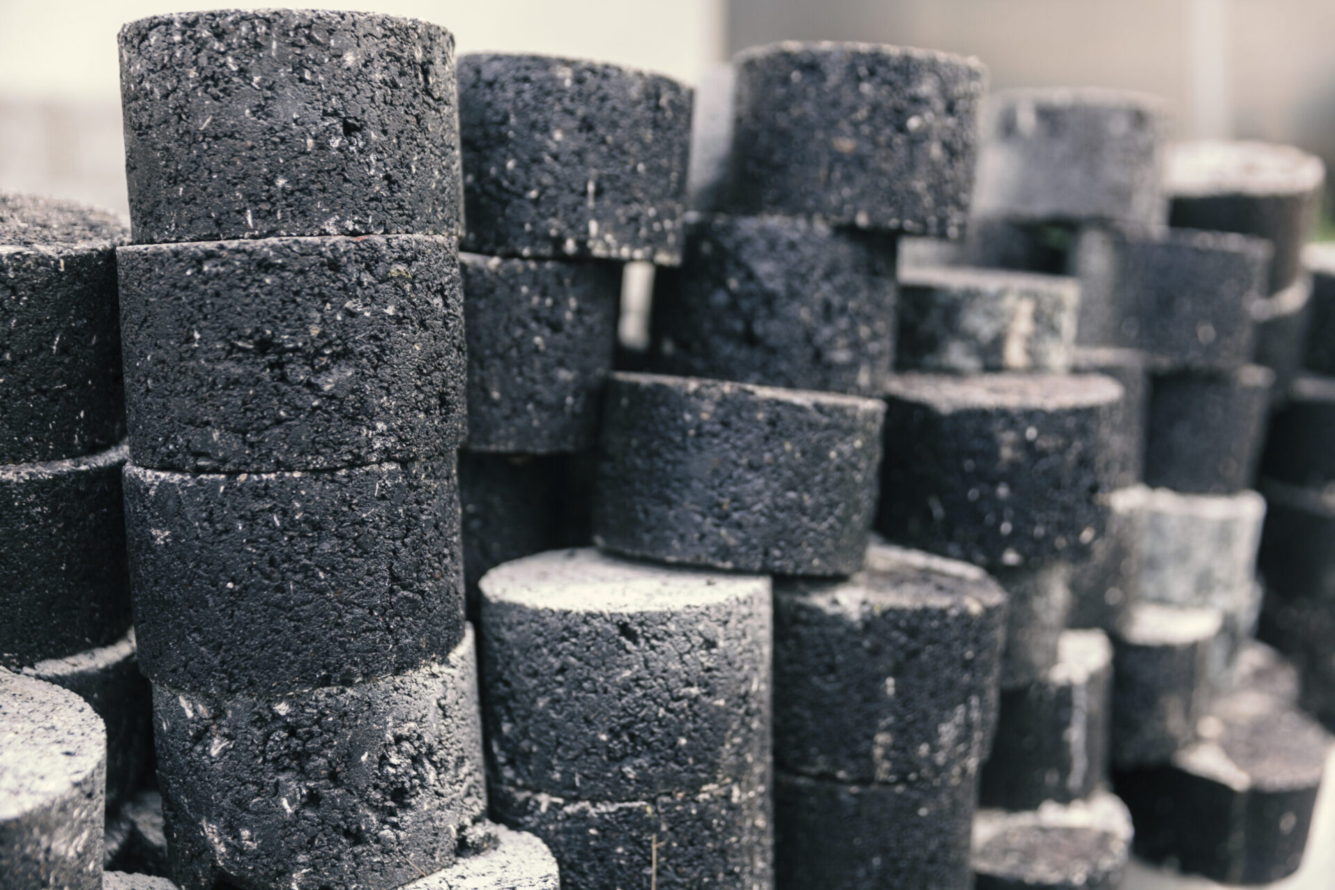 Das Bild zeigt mehrere zylindrische Kernbohrproben aus Asphalt, die aus Straßen entnommen wurden. Die Proben sind in mehreren Stapeln angeordnet und weisen eine raue Oberfläche sowie unterschiedliche Grautöne bis Schwarz auf.