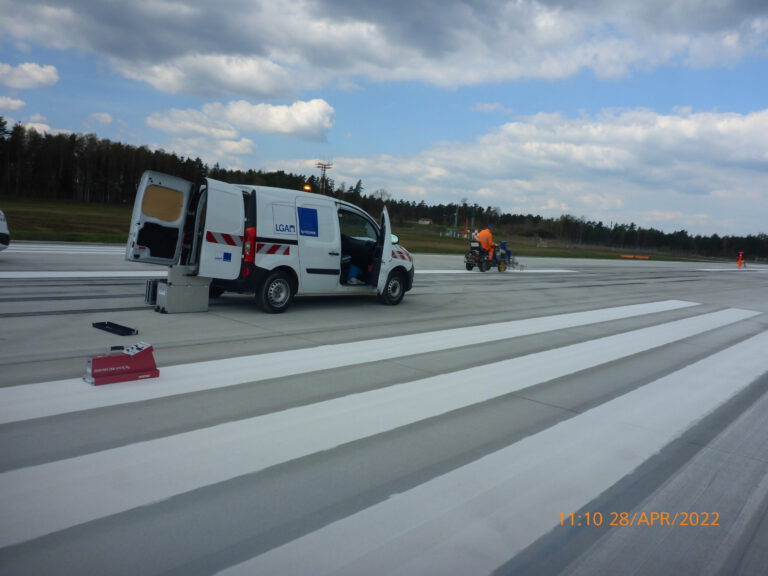 Das Bild zeigt eine Szene auf einer Start- und Landebahn eines Flughafens. Ein weißer Transporter mit geöffneten Türen steht auf der Bahn, daneben arbeitet eine Person in orangefarbener Kleidung an einem Gerät, vermutlich für Markierungs- oder Wartungsarbeiten. Im Hintergrund sind Bäume und ein teilweise bewölkter Himmel zu sehen.