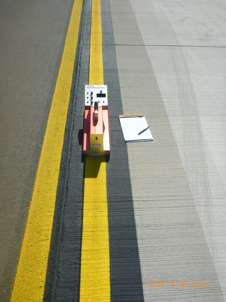Das Bild zeigt einen Straßenabschnitt mit einer gelben Linie am Rand. Auf der Linie steht ein orangefarbenes Gerät, vermutlich ein Mess- oder Vermessungsinstrument. Daneben liegt ein Klemmbrett mit einem Blatt Papier und einem Stift.