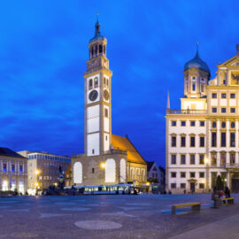 Abendliche Panoramaaufnahme des Augsburger Rathausplatzes mit dem Renaissance-Rathaus in der Bildmitte und dem Perlachturm links daneben, umgeben von beleuchteten Gebäuden und Geschäften.