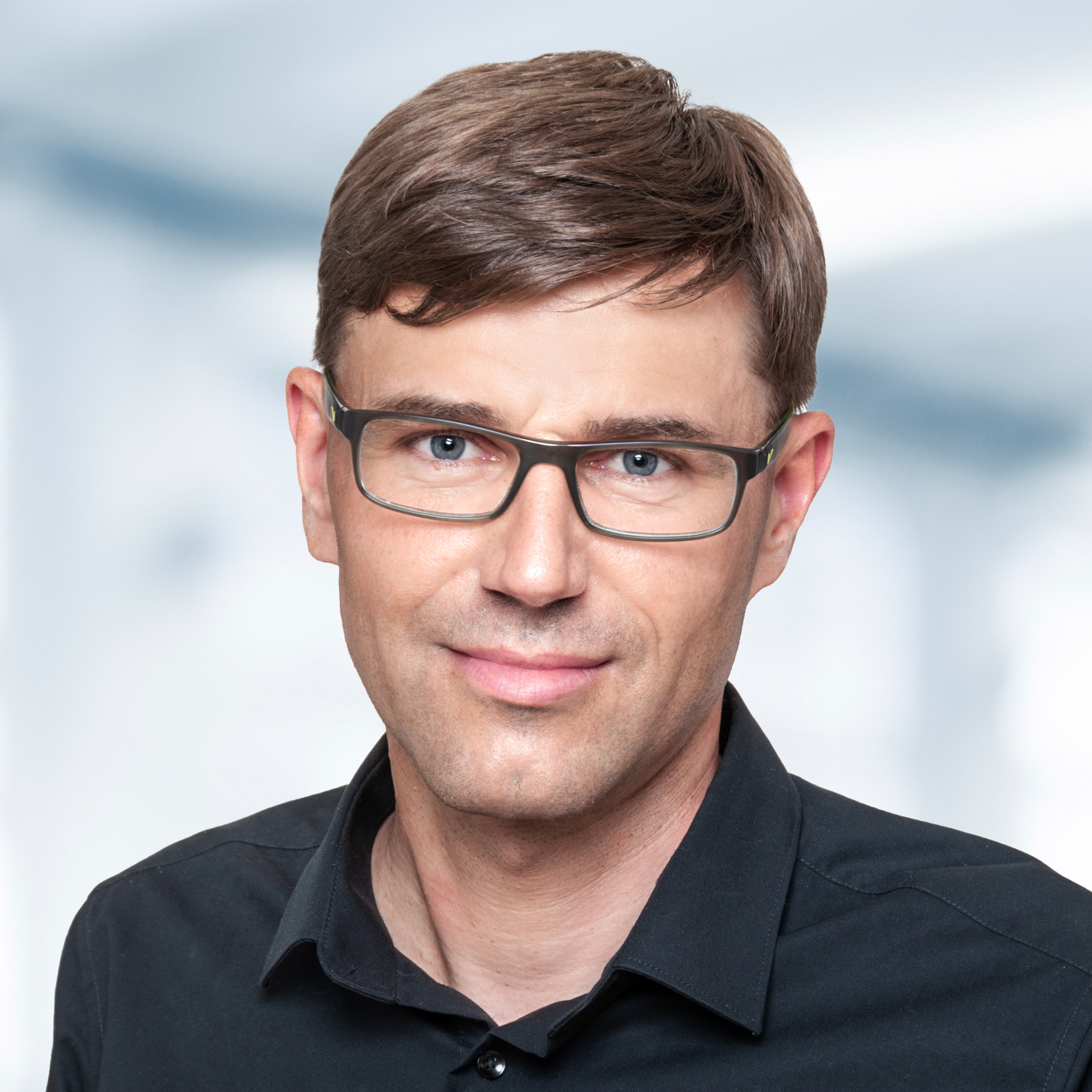 Stefan Baumann trägt ein schwarzes Hemd mit Kragen. Der Hintergrund ist unscharf und zeigt ein Büro.