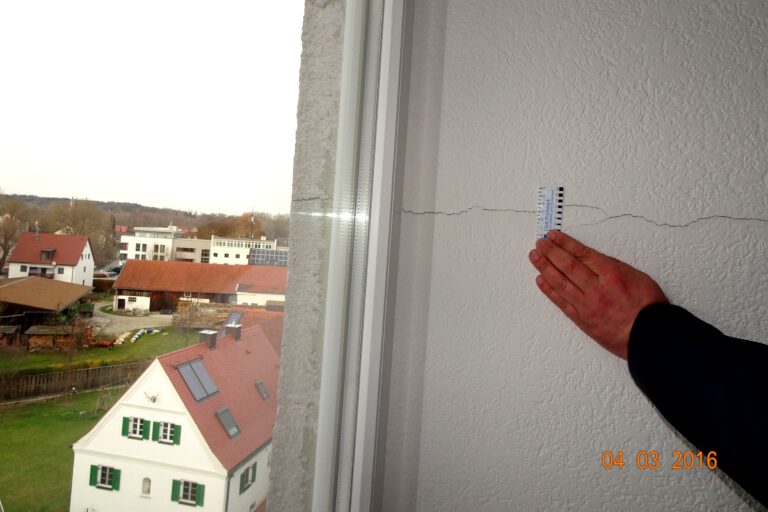 Das Bild zeigt eine Hand, die ein Lineal an eine Wand mit einem sichtbaren Riss hält, um dessen Breite zu messen. Im Hintergrund sind mehrere Häuser und Gebäude zu sehen, einige davon mit Solarpaneelen auf den Dächern.