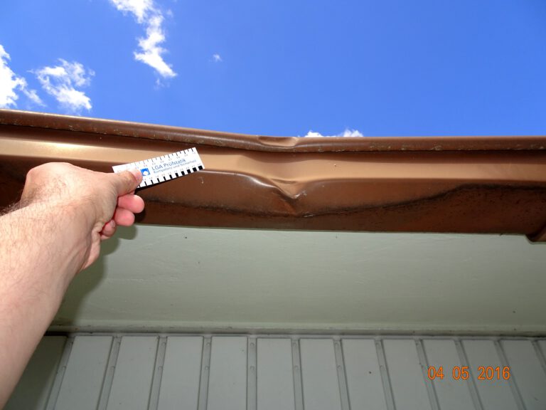 Das Bild zeigt eine Nahaufnahme einer beschädigten braunen Dachrinne am Rand eines Daches. Eine Hand hält ein Lineal neben die Delle in der Rinne, vermutlich um das Ausmaß des Schadens zu messen. Im Hintergrund ist ein teilweise bewölkter Himmel zu sehen.