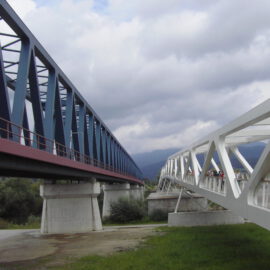 Das Bild zeigt zwei parallel verlaufende Brücken. Links befindet sich eine blaue Stahlfachwerkbrücke mit rotem Unterbau, die von Betonpfeilern getragen wird. Rechts verläuft eine weiße Fußgängerbrücke mit offener Gitterstruktur, auf der mehrere Personen zu sehen sind.