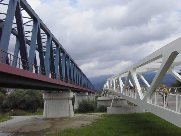 Das Bild zeigt zwei parallel verlaufende Brücken. Links befindet sich eine blaue Stahlfachwerkbrücke mit rotem Unterbau, die von Betonpfeilern getragen wird. Rechts verläuft eine weiße Fußgängerbrücke mit offener Gitterstruktur, auf der mehrere Personen zu sehen sind.
