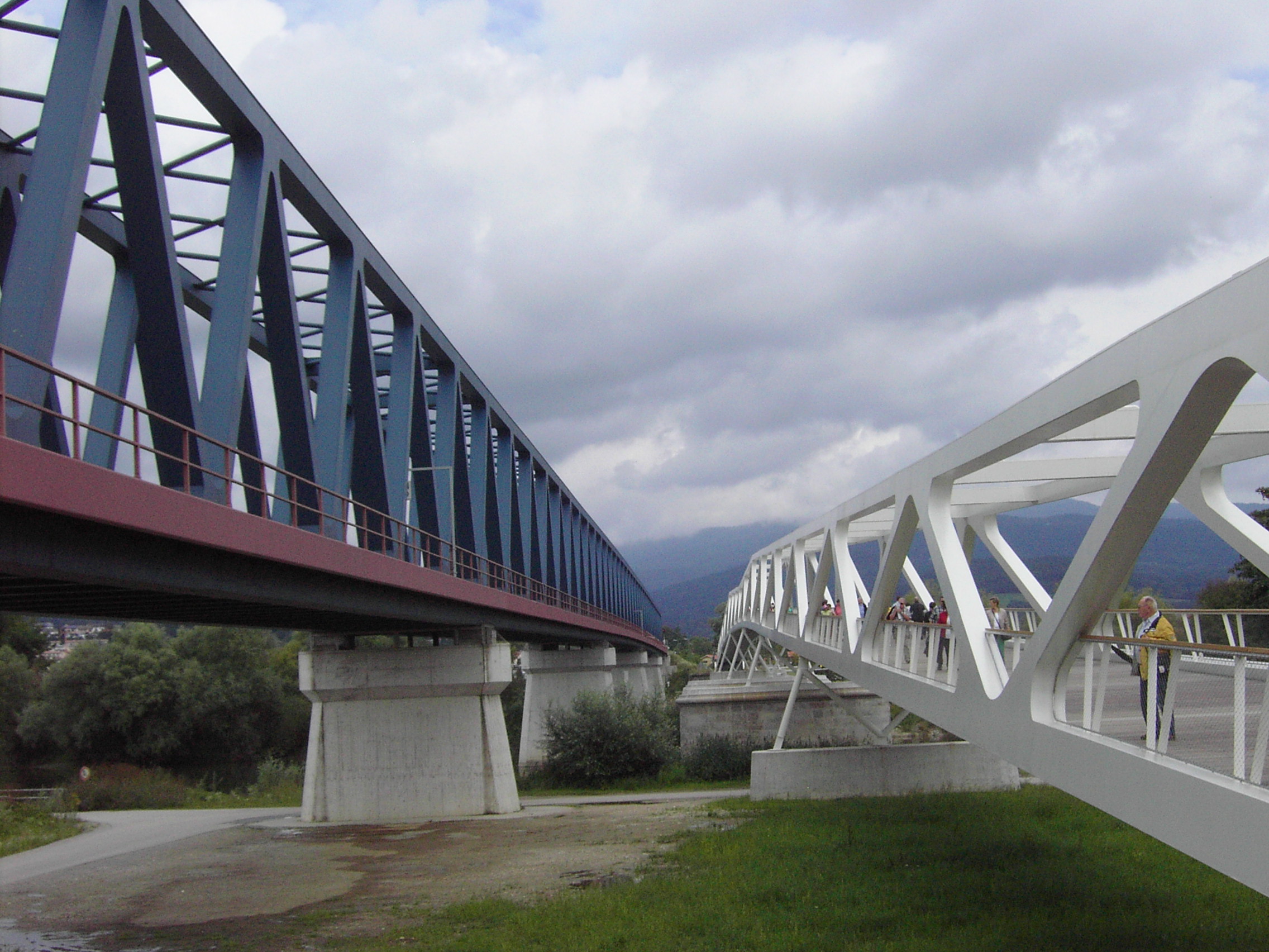 Das Bild zeigt zwei parallel verlaufende Brücken. Links befindet sich eine blaue Stahlfachwerkbrücke mit rotem Unterbau, die von Betonpfeilern getragen wird. Rechts verläuft eine weiße Fußgängerbrücke mit offener Gitterstruktur, auf der mehrere Personen zu sehen sind.