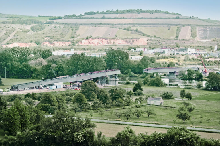 Das Bild zeigt eine teilweise errichtete Brücke, die über einen Fluss in einer ländlichen Gegend führt. Mehrere Abschnitte der Brücke sind bereits vorhanden und werden von provisorischen Konstruktionen gestützt. In der Nähe der Brücke sind Baustellenfahrzeuge und Geräte zu sehen, die auf laufende Arbeiten hinweisen.