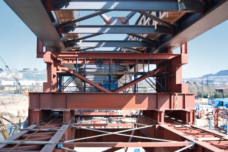 Das Bild zeigt eine Baustelle mit dem Fokus auf das Stahlgerüst einer Brücke. Die Perspektive ist von unten, sodass man durch die Träger und Stützen blickt, die in einem gitterartigen Muster angeordnet sind. Im Hintergrund sind Baumaterialien, Maschinen und einige Arbeiter zu sehen, dahinter erstreckt sich eine hügelige Landschaft.