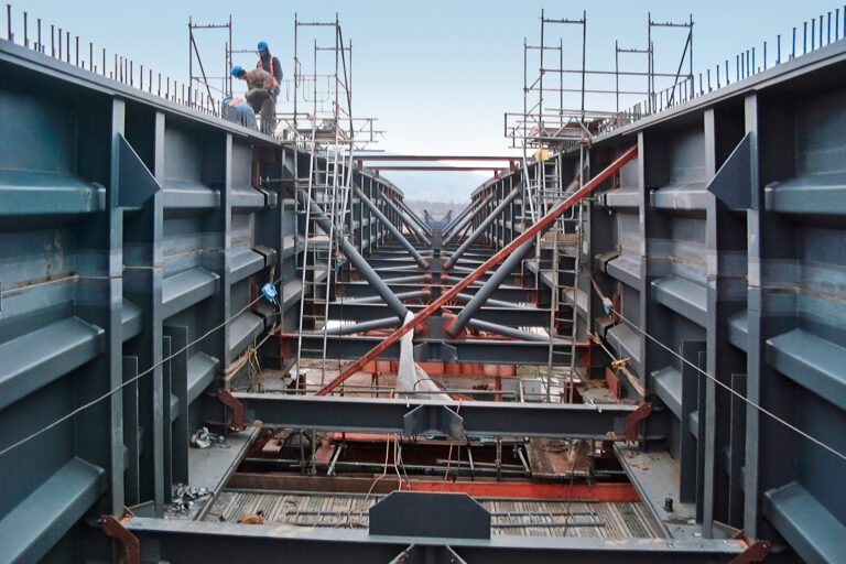 Das Bild zeigt eine Baustelle mit großen Stahlkonstruktionen und Gerüsten. Auf dem Gerüst befinden sich mehrere Arbeiter, und es sind verschiedene Träger und Stützen zu sehen. Die Szene zeigt den Bau einer Brücke bei klarem Tageslicht.