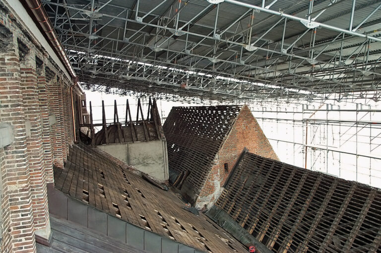 Das Bild zeigt mehrere alte, beschädigte Dächer von Ziegelgebäuden, die sich unter einer großen Metallgerüstkonstruktion befinden. Die Dächer sind unterschiedlich stark beschädigt, mit fehlenden Ziegeln und freiliegenden Holzbalken. Das Gerüst ist Teil eines Sanierungs- oder Bauprojekts und dient dem Schutz und der Stabilisierung der Gebäude.