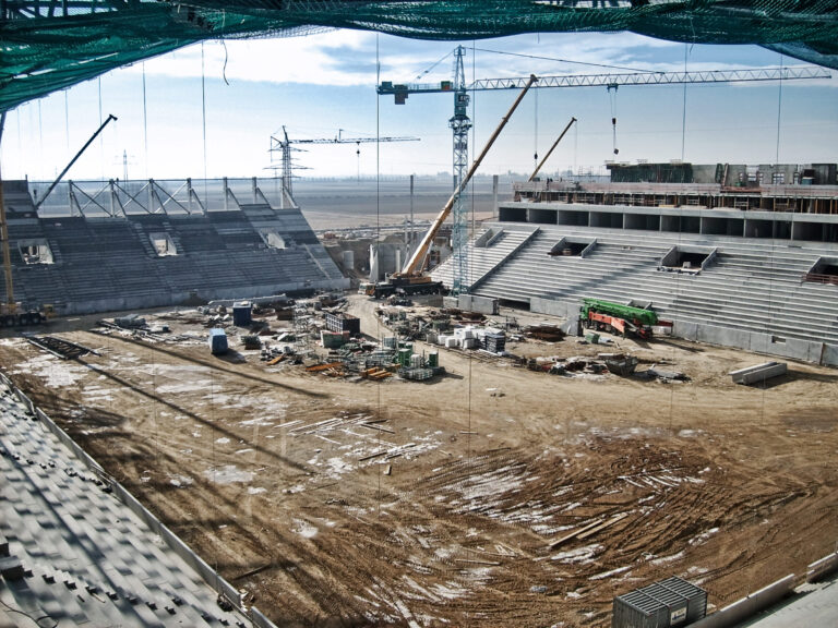 Auf dem Bild ist eine große Baustelle eines Stadions zu sehen. Mehrere Kräne und verschiedene Baumaschinen befinden sich auf dem Gelände, während die Tribünenbereiche teilweise fertiggestellt sind. Im Innenraum des Stadions liegen Baumaterialien und Geräte verteilt.