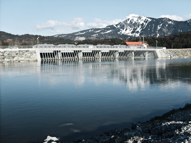 Das Bild zeigt ein Stauwerk mit mehreren Schleusentoren, die zur Regulierung des Wasserflusses dienen. Im Vordergrund befindet sich ein großer Wasserkörper, im Hintergrund sind schneebedeckte Berge zu sehen. Rechts neben dem Stauwerk steht ein Gebäude mit orangefarbenem Dach, umgeben von Bäumen und Vegetation.