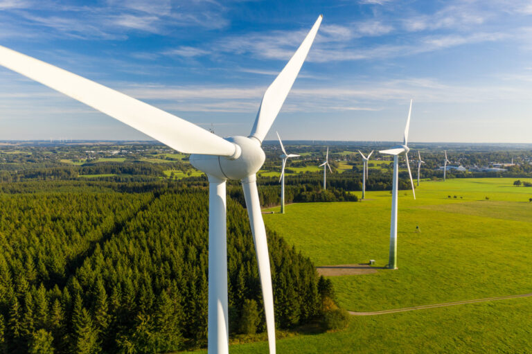 Das Bild zeigt einen Windpark mit mehreren großen Windrädern, die in einer grünen, grasbewachsenen Landschaft stehen. Die Windräder sind über das Feld verteilt, im Hintergrund sind einige Bäume zu sehen. Der Himmel ist klar und blau, was auf einen sonnigen Tag hinweist.