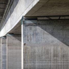 Das Bild zeigt eine Nahaufnahme der Unterseite einer Betonbrücke mit großen Stützpfeilern und Trägern. Die Oberflächen weisen sichtbare Strukturen und Schatten auf. Im Hintergrund ist grüne Vegetation zu erkennen, was auf eine ländliche oder naturnahe Umgebung hinweist.