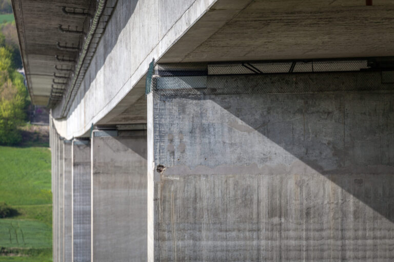 Das Bild zeigt eine Nahaufnahme der Unterseite einer Betonbrücke mit großen Stützpfeilern und Trägern. Die Oberflächen weisen sichtbare Strukturen und Schatten auf. Im Hintergrund ist grüne Vegetation zu erkennen, was auf eine ländliche oder naturnahe Umgebung hinweist.
