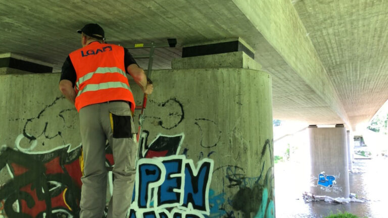 Das Bild zeigt eine Person mit Warnweste, die unter einer Betonbrücke arbeitet. Die Person überprüft einen Brückenpfeiler. Im Hintergrund ist ein weiterer Brückenpfeiler mit Graffiti auf der gegenüberliegenden Wasserseite zu sehen.