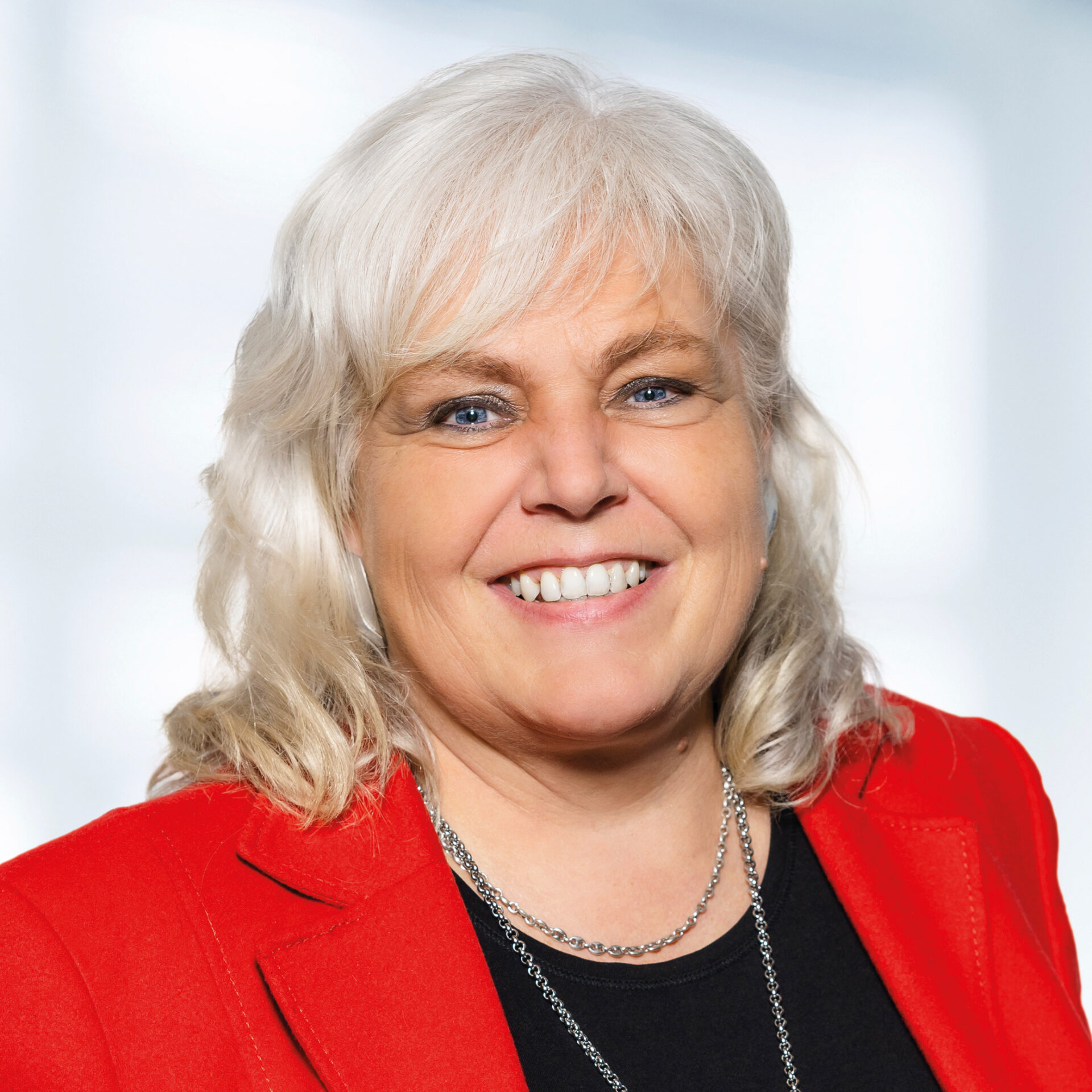 Eine Person mit hellen Haaren trägt einen roten Blazer über einem schwarzen Oberteil. Sie steht in einem Innenraum mit natürlichem Lichteinfall und blickt lächelnd in die Kamera. Der Hintergrund ist unscharf und zeigt Fenster, durch die Tageslicht fällt.