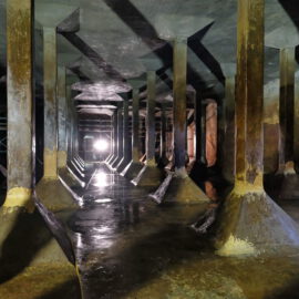 Das Bild zeigt eine unterirdische Struktur mit mehreren Betonsäulen, die die Decke tragen. Der Boden ist nass und spiegelt das Licht einer hellen Lichtquelle am Ende des Raumes wider. Die Säulen sind in Reihen angeordnet und erzeugen ein symmetrisches, sich wiederholendes Muster.