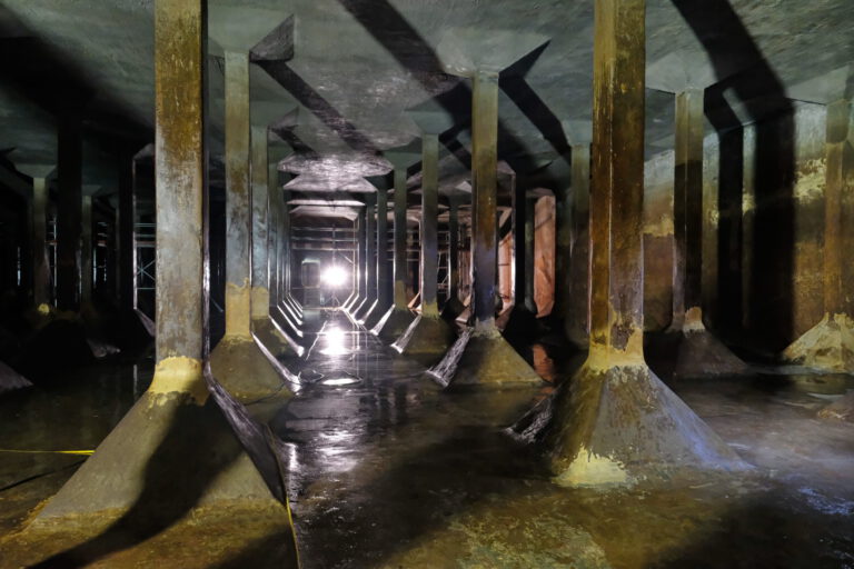 Das Bild zeigt eine unterirdische Struktur mit mehreren Betonsäulen, die die Decke tragen. Der Boden ist nass und spiegelt das Licht einer hellen Lichtquelle am Ende des Raumes wider. Die Säulen sind in Reihen angeordnet und erzeugen ein symmetrisches, sich wiederholendes Muster.