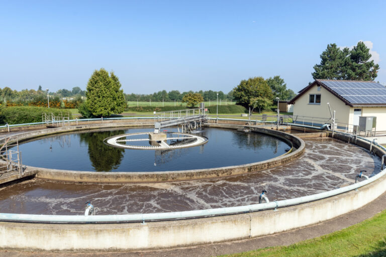 Das Bild zeigt ein rundes Betonbecken einer Kläranlage, ein Absetz- oder Klärbecken. In der Mitte befindet sich eine mechanische Vorrichtung zur Entfernung von Feststoffen aus dem Wasser. Im Hintergrund steht ein kleines Gebäude mit Solarpaneelen auf dem Dach, umgeben von Bäumen und Grünflächen.