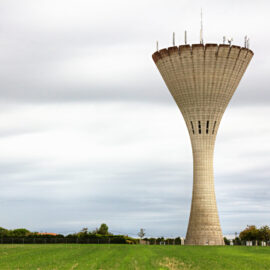 Das Bild zeigt einen großen Betonturm mit ausladender Spitze, der in einem offenen Feld mit grünem Gras steht. Der Himmel ist bewölkt und grau, im Hintergrund sind Bäume, Büsche, ein Zaun und Stromleitungen zu sehen. Auf dem oberen Rand des Turms sind mehrere Antennen oder technische Geräte montiert.