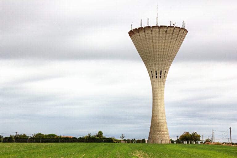 Das Bild zeigt einen großen Betonturm mit ausladender Spitze, der in einem offenen Feld mit grünem Gras steht. Der Himmel ist bewölkt und grau, im Hintergrund sind Bäume, Büsche, ein Zaun und Stromleitungen zu sehen. Auf dem oberen Rand des Turms sind mehrere Antennen oder technische Geräte montiert.