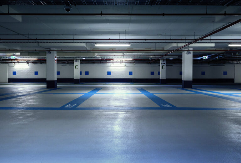 Das Bild zeigt eine leere Tiefgarage mit blauen und weißen Markierungen auf dem Boden zur Kennzeichnung der Parkplätze. Die Decke ist mit verschiedenen Rohrleitungen und Beleuchtungseinrichtungen ausgestattet, und mehrere Betonsäulen sind mit dem Buchstaben C gekennzeichnet. Die Beleuchtung ist künstlich und erzeugt eine leicht gedämpfte Atmosphäre.