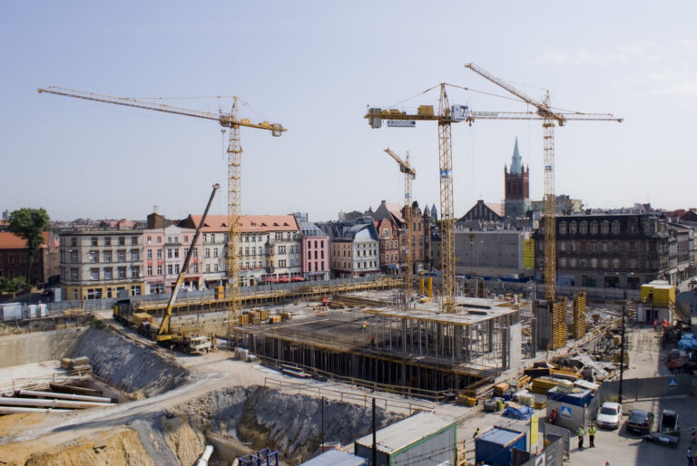 Das Bild zeigt eine Baustelle mit mehreren Kränen und teilweise errichteten Gebäuden. Die Umgebung besteht aus älteren Gebäuden, was auf eine städtische Lage hinweist. Im Hintergrund sind bunte Häuserreihen und ein Kirchturm mit Spitze zu sehen.