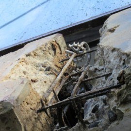 Das Bild zeigt eine Nahaufnahme eines beschädigten Betonbauteils mit freiliegender, verrosteter Bewehrung. Der Beton ist brüchig und teilweise abgeplatzt, wodurch die inneren Metallverstärkungen sichtbar werden. Zwischen den Bruchstellen ist ein Kunststoffelement mit mehreren Speichen zu erkennen.