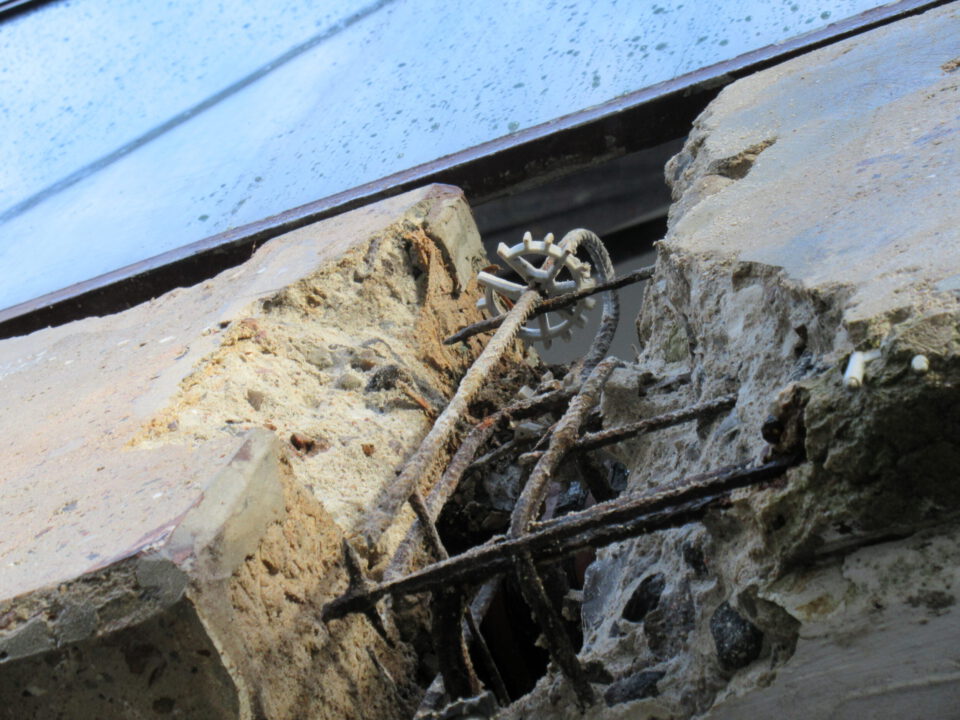 Das Bild zeigt eine Nahaufnahme eines beschädigten Betonbauteils mit freiliegender, verrosteter Bewehrung. Der Beton ist brüchig und teilweise abgeplatzt, wodurch die inneren Metallverstärkungen sichtbar werden. Zwischen den Bruchstellen ist ein Kunststoffelement mit mehreren Speichen zu erkennen.