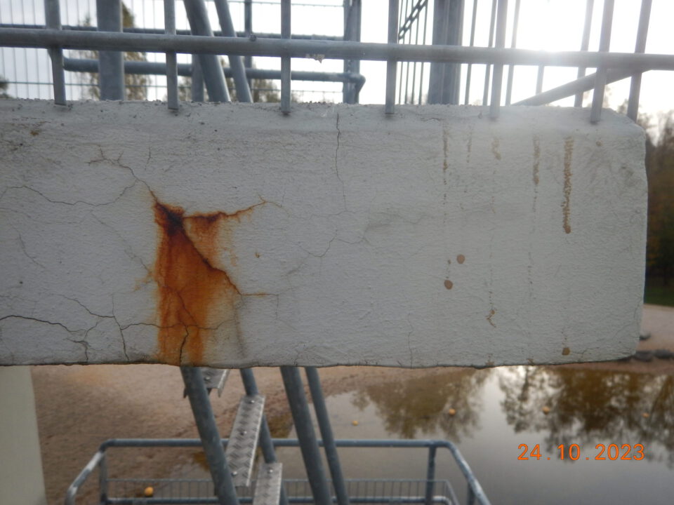 Das Bild zeigt eine Betonfläche mit sichtbaren Schäden und Rostspuren. Mehrere Risse durchziehen die Oberfläche, und aus dem oberen linken Bereich zieht sich ein deutlicher Rostfleck nach unten, was auf eine Korrosion der Bewehrung hinweist.