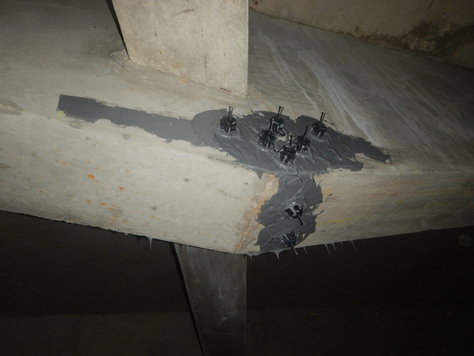 Das Bild zeigt einen Betonträger mit sichtbaren Rissen, die durch ein Epoxidharz-Injektionsverfahren instandgesetzt wurden. Die Risse sind mit grauem Harz versiegelt, und entlang der Risslinien sind mehrere kleine schwarze Injektionsstellen angebracht. Das Harz wurde großzügig aufgetragen und bedeckt sowohl die Risse als auch angrenzende Flächen.