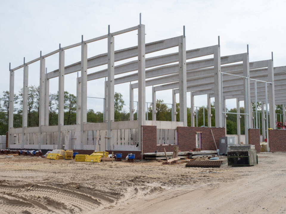 Das Bild zeigt die Baustelle einer neuen Produktionshalle mit einem Skelett aus Stahlbeton. Im unteren Bereich ist eine Verblendung aus Mauerwerk zu sehen. Auf dem Gelände liegen verschiedene Baumaterialien und Baugeräte.