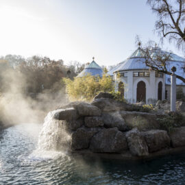 Das Bild zeigt eine ruhige Außenaufnahme mit einem kleinen Wasserfall, der über Felsen in ein Wasserbecken fließt. Im Hintergrund stehen weiße Gebäude mit blauen Dächern und Rundbögen, umgeben von Bäumen und Pflanzen. Ein leichter Nebel liegt über der Szene, vermutlich durch die Gischt des Wassers.