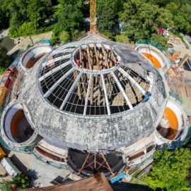 Das Bild zeigt eine Luftaufnahme einer Baustelle mit einem großen, runden Gebäude, das sich im Umbau oder Bau befindet. Die zentrale Kuppelstruktur ist von Gerüsten und freiliegenden Trägern umgeben, daneben befinden sich mehrere kleinere runde Bauwerke. Um das Gelände herum sind Bäume und Grünflächen zu sehen.