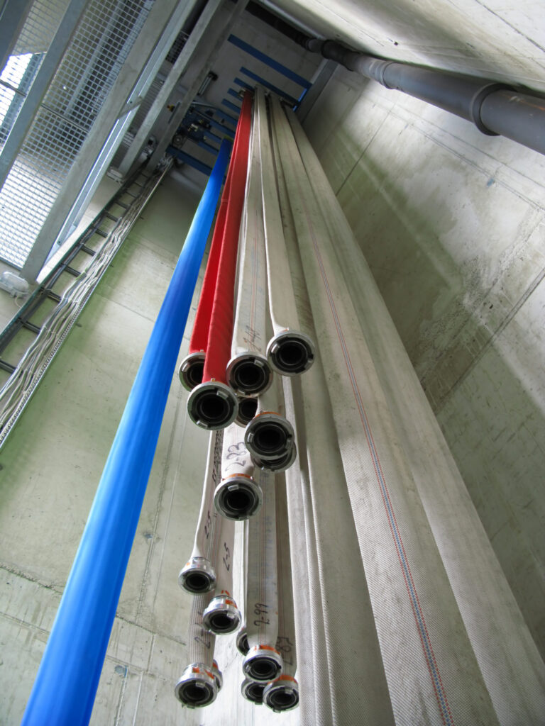 Das Bild zeigt mehrere lange Schläuche, die senkrecht in einem Lagerbereich hängen. Die Schläuche sind in verschiedenen Farben wie Blau, Rot und Weiß zu sehen und unten an Metallkupplungen befestigt. Sie sind in parallelen Reihen angeordnet, im Hintergrund sind Betonwände und Metallstrukturen zu erkennen.