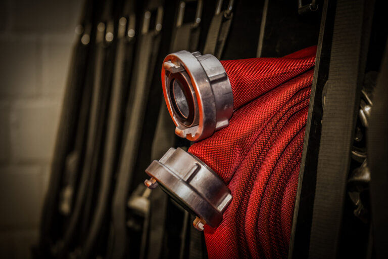 Das Bild zeigt eine Nahaufnahme eines roten Feuerwehrschlauchs mit Metallkupplungen, der ordentlich auf einem Gestell aufgerollt ist. Im Hintergrund befinden sich weitere ähnliche Schläuche und Ausrüstungsgegenstände, was auf eine Feuerwache oder eine Notfallausrüstung hinweist.