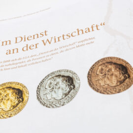 Das Bild zeigt eine Seite aus einer Publikation mit dem Titel „Im Dienst an der Wirtschaft“. Unter dem Titel steht ein erklärender Text zur Bedeutung dieses Mottos. Darunter sind drei Medaillen abgebildet – von links nach rechts in Gold, Silber und Bronze – jeweils mit zwei Profilporträts und umlaufender Beschriftung.