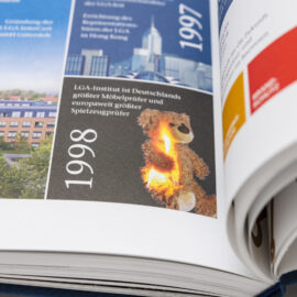 Das Bild zeigt ein geöffnetes Buch mit einer Zeitleiste, auf der die Jahre 1997 und 1998 hervorgehoben sind. Im Abschnitt zu 1998 steht, dass das LGA-Institut Deutschlands größter Möbelprüfer und europaweit größter Spielzeugprüfer ist. Daneben ist ein brennender Teddybär abgebildet.