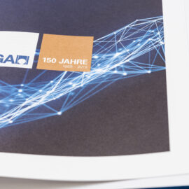 Das Bild zeigt eine Seite aus einem Buch oder Magazin mit dunklem Hintergrund und einem abstrakten Muster aus blauen Linien und Punkten. In der Mitte befinden sich zwei farbige Quadrate: ein weißes Quadrat mit der Aufschrift „LGA“ und geometrischem Logo sowie ein bronzenes Quadrat mit der Aufschrift „150 JAHRE 1869 – 2019“.