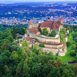 Luftaufnahme der Veste Coburg, einer mittelalterlichen Festung mit roten Ziegeldächern und Steinmauern, umgeben von dichten Wäldern und mit Blick auf die Stadt Coburg im Hintergrund.